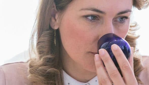 Woman with curly hair holding a blue glass container to her nose while smelling its contents. - Olive Oil Times