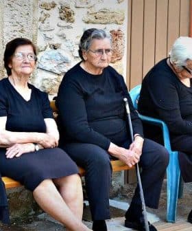 Six elderly women dressed in black and gray sitting on a bench in a casual setting. - Olive Oil Times