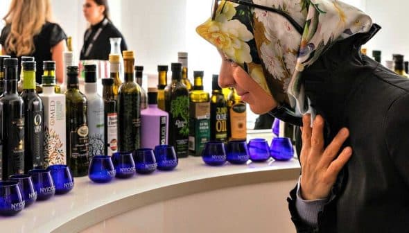 Woman wearing a floral headscarf examining various bottles of olive oil on display. - Olive Oil Times