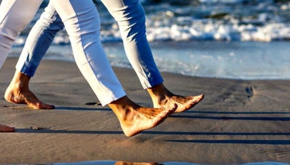 Two individuals walking barefoot along the shoreline with waves in the background. - Olive Oil Times
