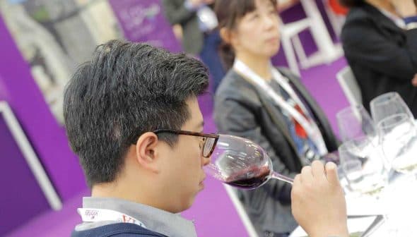 Individual holding a wine glass and tasting red wine at a gathering with others in the background. - Olive Oil Times