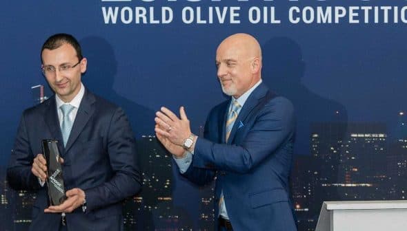 Two men at the World Olive Oil Competition award ceremony, one holding a trophy and the other applauding. - Olive Oil Times