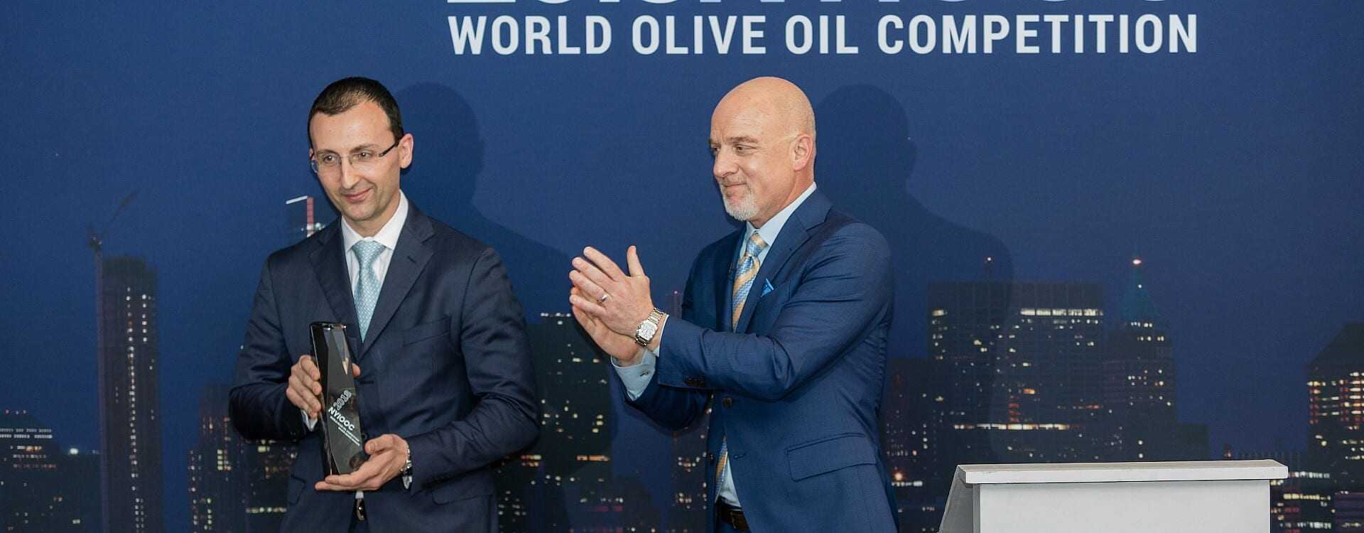 Two men at the World Olive Oil Competition award ceremony, one holding a trophy and the other applauding. - Olive Oil Times