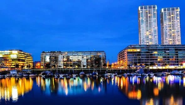Night view of a city skyline with buildings and boats reflected in the water. - Olive Oil Times