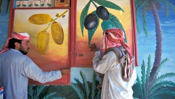 Two men in traditional attire examining wall murals depicting olives and dates. - Olive Oil Times