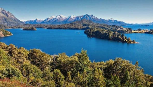 A panoramic view of a lake surrounded by mountains and forests under a clear blue sky. - Olive Oil Times