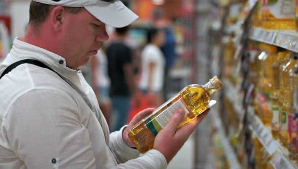 Individual inspecting a bottle of cooking oil while standing in a grocery store aisle. - Olive Oil Times