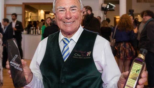 Man in a green vest holding an award and a bottle of olive oil at an event. - Olive Oil Times