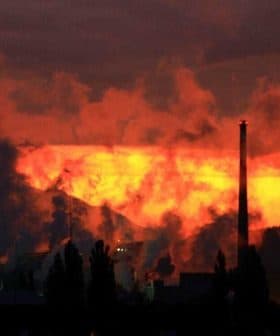 Silhouetted industrial structures emitting smoke against a vibrant sunset sky. - Olive Oil Times