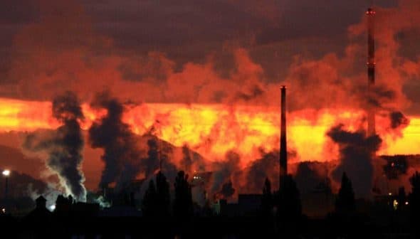 Silhouetted industrial structures emitting smoke against a vibrant sunset sky. - Olive Oil Times