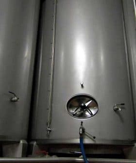 Three stainless steel fermentation tanks with circular openings and valves. - Olive Oil Times