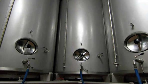 Three stainless steel fermentation tanks with circular openings and valves. - Olive Oil Times