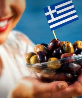 A woman smiling while holding a bowl of assorted olives topped with a small Greek flag. - Olive Oil Times