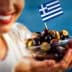 A woman smiling while holding a bowl of assorted olives topped with a small Greek flag. - Olive Oil Times