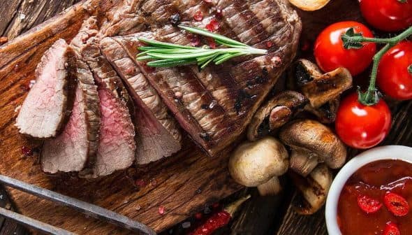 Sliced grilled steak on a wooden cutting board with tomatoes, mushrooms, and sauce. - Olive Oil Times