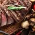 Sliced grilled steak on a wooden cutting board with tomatoes, mushrooms, and sauce. - Olive Oil Times