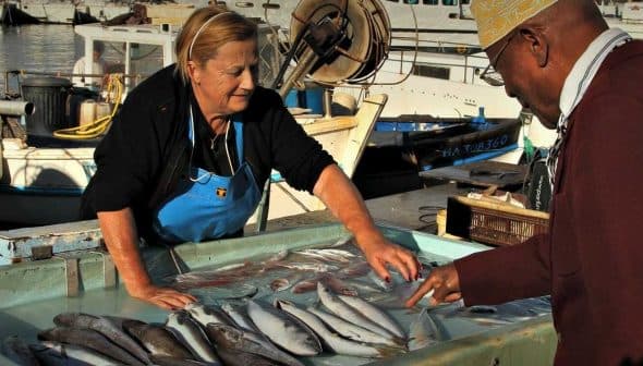 A woman in an apron selling fish at a market while a man points at the fish on display. - Olive Oil Times