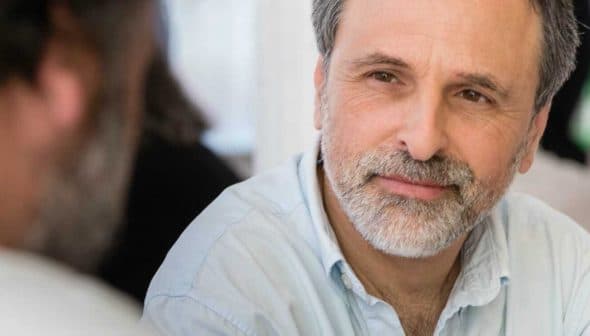 A man with a beard wearing a light blue shirt, engaged in conversation with another person. - Olive Oil Times