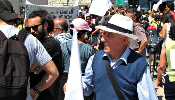 An older man wearing a white hat and holding a small flag at a crowded public event. - Olive Oil Times