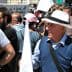 An older man wearing a white hat and holding a small flag at a crowded public event. - Olive Oil Times