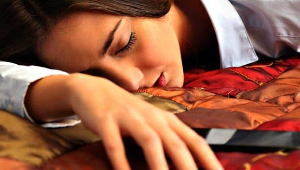 A woman resting her head on a bed with her arm extended towards a smartphone. - Olive Oil Times