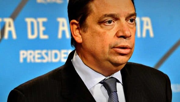 Man in a dark suit and tie speaking at a press conference with a blue background. - Olive Oil Times