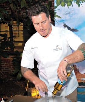 Chef in a white uniform mixing ingredients in a bowl at an outdoor event with a backdrop. - Olive Oil Times