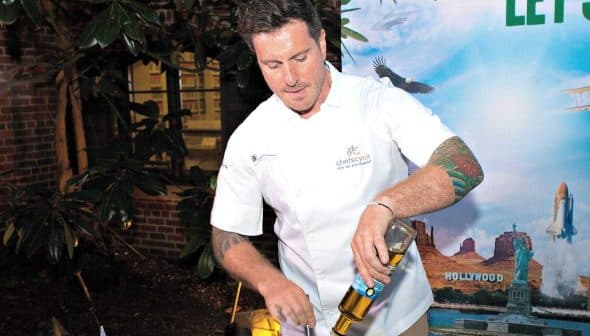 Chef in a white uniform mixing ingredients in a bowl at an outdoor event with a backdrop. - Olive Oil Times