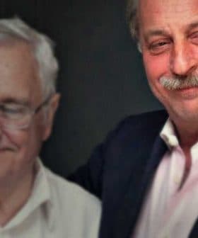 Two men standing close together, smiling for a photograph in a dimly lit setting. - Olive Oil Times