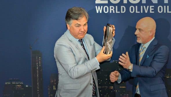 Two men at an award ceremony, one presenting a bottle to the other in front of a city skyline backdrop. - Olive Oil Times