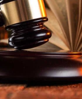 A wooden gavel resting on a round base next to an open book on a wooden table. - Olive Oil Times