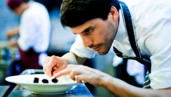 Chef carefully plating food on a white plate in a professional kitchen setting. - Olive Oil Times