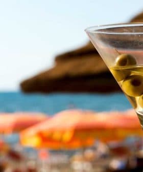 A martini glass filled with a clear liquid and three green olives, with a beach background. - Olive Oil Times