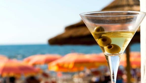 A martini glass filled with a clear liquid and three green olives, with a beach background. - Olive Oil Times