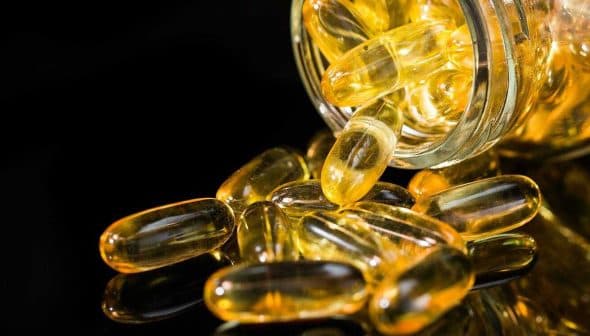 A glass jar tipped over with yellow gel capsules spilling out onto a reflective surface. - Olive Oil Times