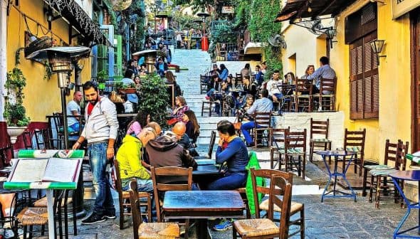 Outdoor dining area featuring wooden tables and chairs in a bustling setting with patrons. - Olive Oil Times