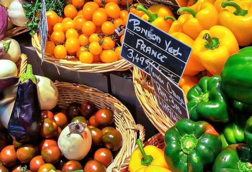 A display of various fresh vegetables and fruits including peppers, tomatoes, and eggplants in baskets. - Olive Oil Times