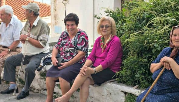 Five elderly individuals sitting on a stone wall in a village setting, with greenery in the background. - Olive Oil Times