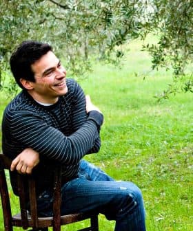 Man with dark hair sitting on a wooden chair, smiling in an olive grove. - Olive Oil Times