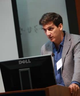 A man in a gray blazer speaking at a podium with a Dell monitor during a presentation. - Olive Oil Times