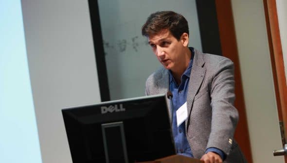 A man in a gray blazer speaking at a podium with a Dell monitor during a presentation. - Olive Oil Times