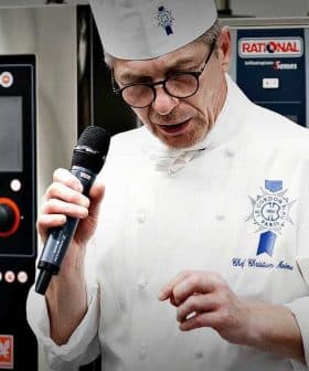 Chef in a white uniform and hat speaking into a microphone at a culinary event. - Olive Oil Times