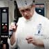Chef in a white uniform and hat speaking into a microphone at a culinary event. - Olive Oil Times