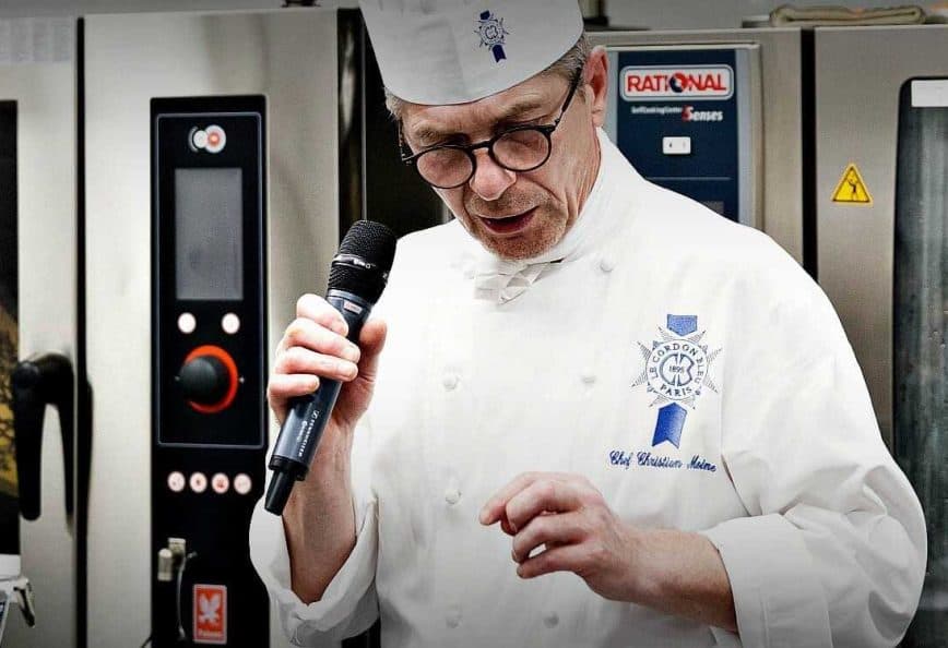Chef in a white uniform and hat speaking into a microphone at a culinary event. - Olive Oil Times