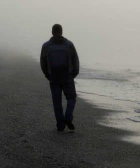 Individual walking alone on a foggy beach with gentle waves lapping at the shore. - Olive Oil Times