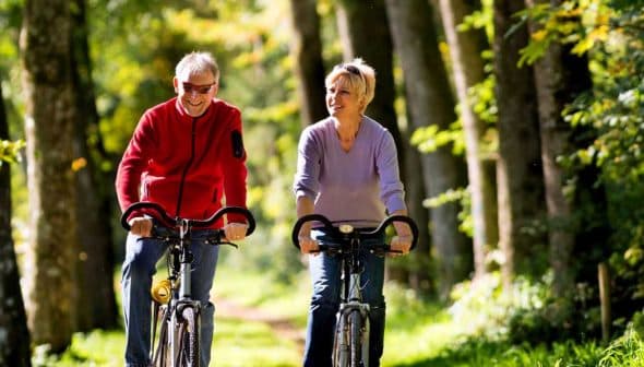 A man and woman riding bicycles side by side on a tree-lined path during daylight. - Olive Oil Times