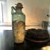 An ancient glass bottle with a cork stopper and a dark round object beside it on a display table. - Olive Oil Times