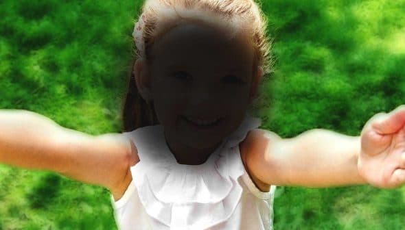 A child with outstretched arms smiling at the camera, wearing a white top with ruffles. - Olive Oil Times