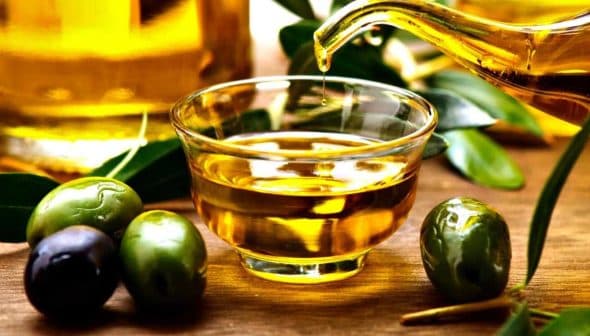 Glass container pouring olive oil into a small glass bowl with olives beside it. - Olive Oil Times