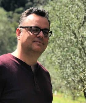 Man wearing glasses standing in an olive grove with trees in the background. - Olive Oil Times